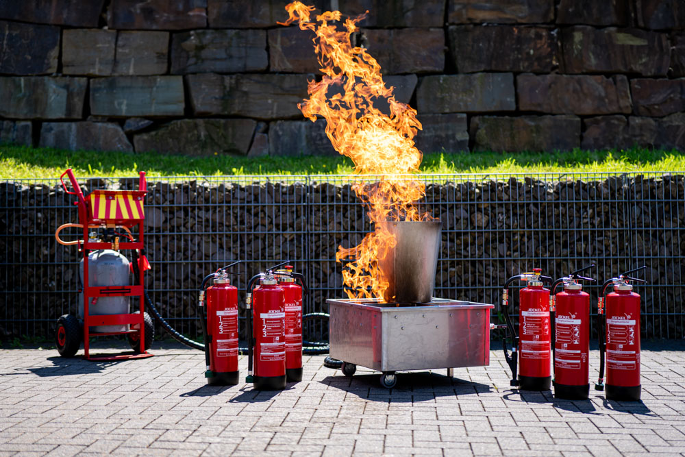 Aufbau einer Brandschutzübung mit mehreren Feuerlöschern und einem brennenden Behälter im Freien.